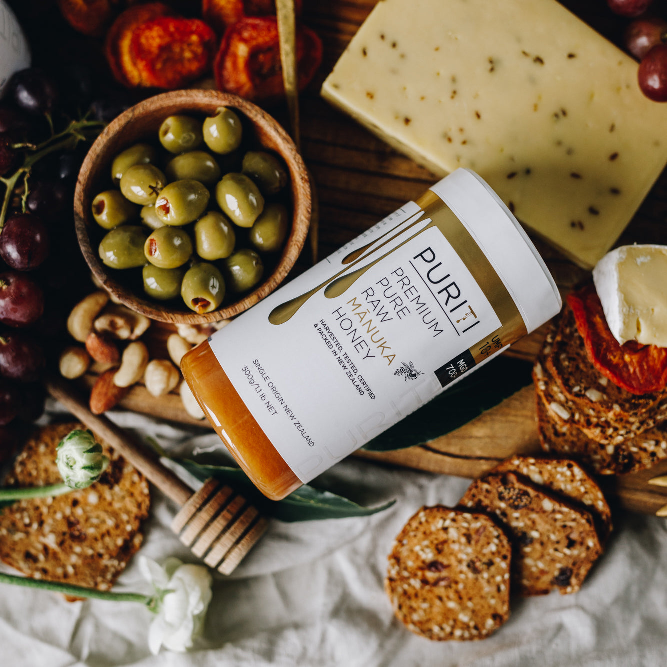 PURITI Premium Raw Manuka Honey UMF 18+ | MGO 700, olives, nuts, and crackers on a cutting board. (Brand: novita SG)