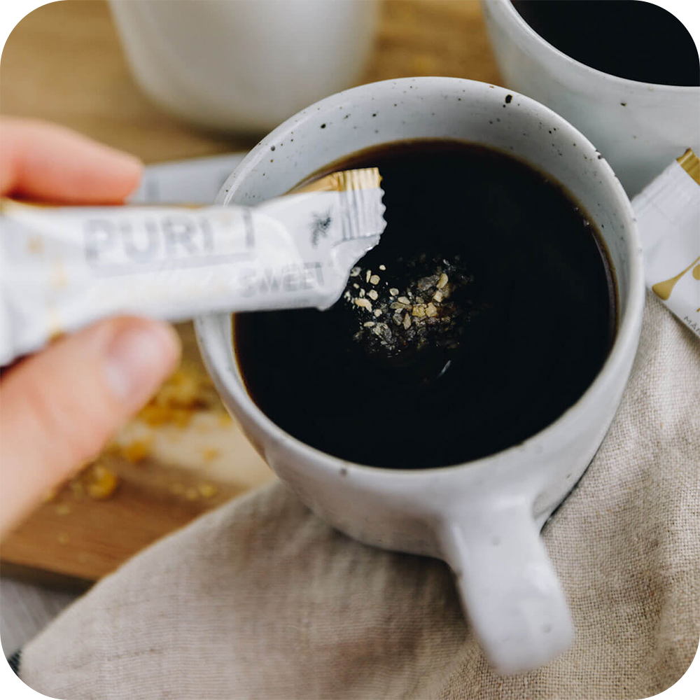 A person is holding a bar of PURITI Manuka Honey Crystals UMF 5+ | MGO 100 from the brand novita SG in a cup of coffee.