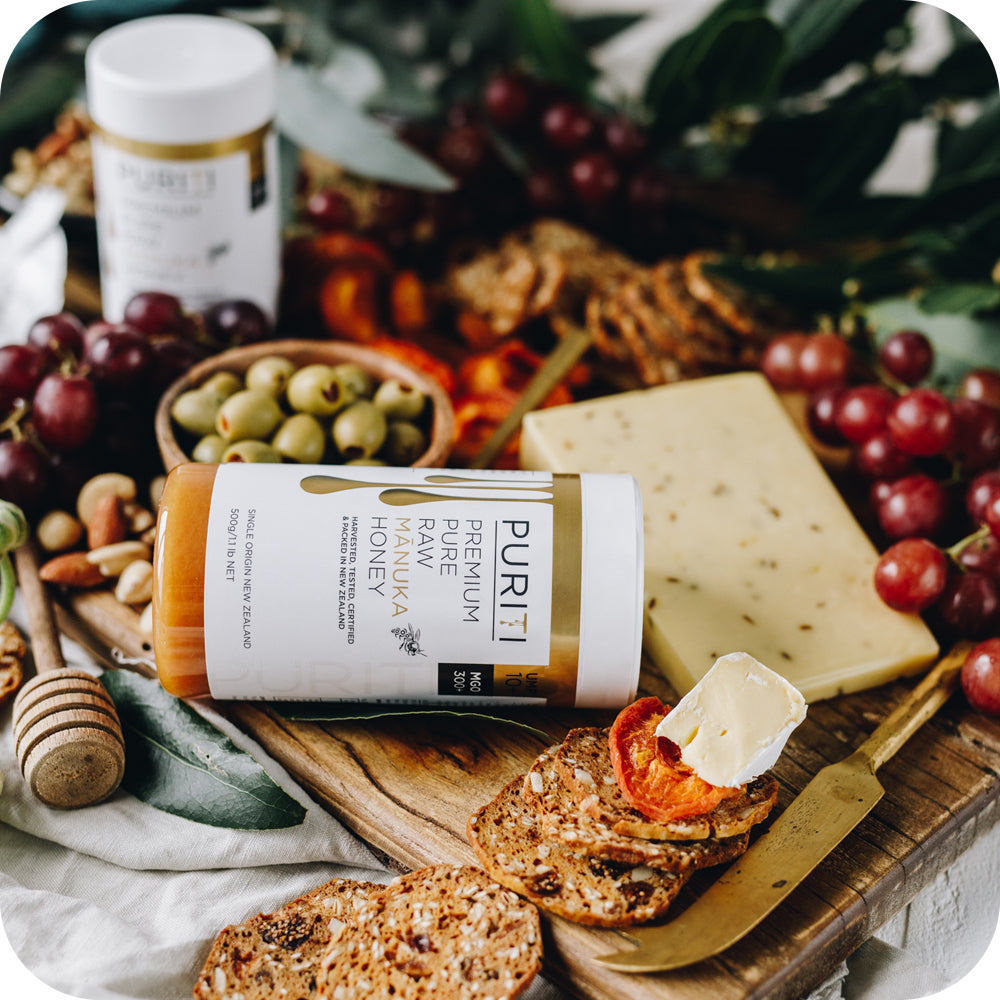 A high-end cutting board with grapes, nuts, and a bottle of novita SG PURITI Premium Raw Manuka Honey UMF 10+ | MGO 300 (6 Bottles Family Pack).