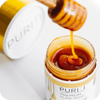 A person is pouring PURITI Premium Raw Manuka Honey UMF 18+ | MGO 700 from novita SG into a jar.